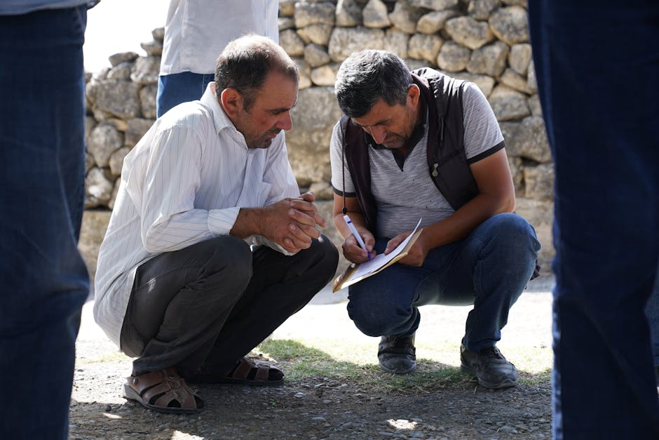 Two men squatting and analyzing paperwork outdoors in a sunny setting.